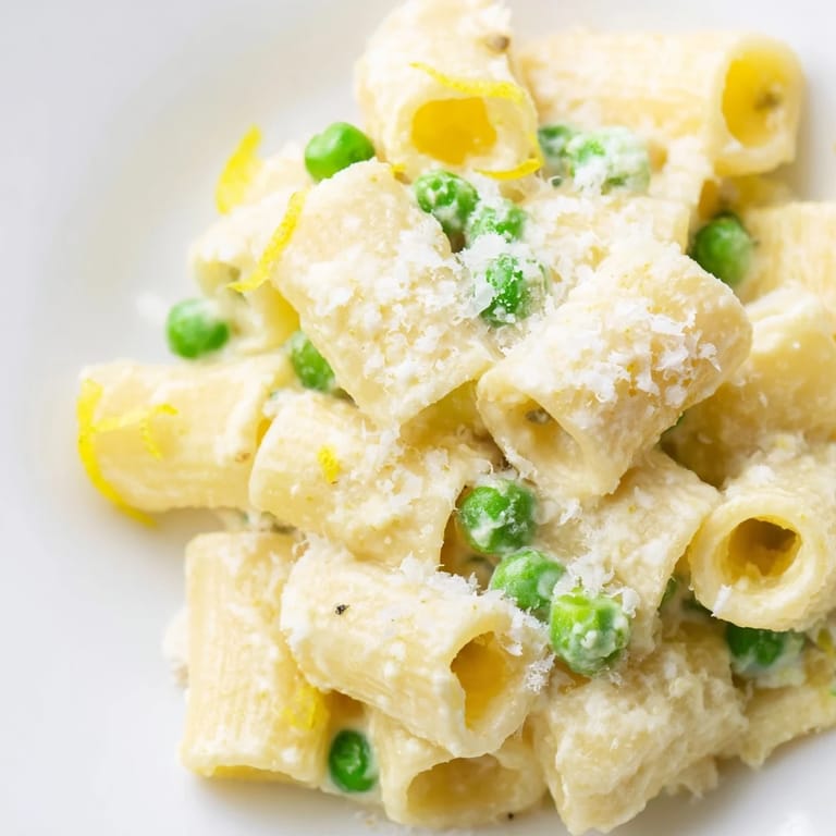 Shiny rigatoni pasta tossed with peas and creamy ricotta sauce, garnished with lemon zest and a drizzle of olive oil.